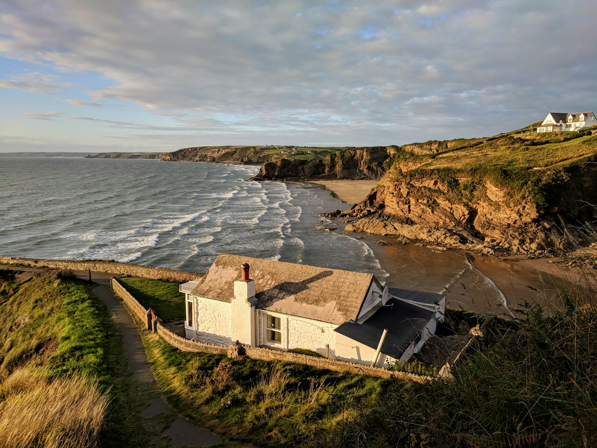 A white-painted clifftop cottage at sunset, overlooking a sandy cove and breaking waves